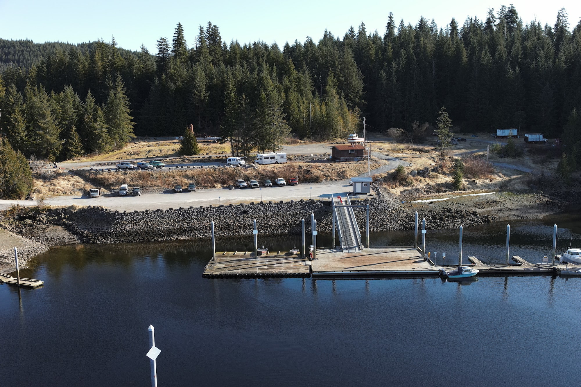 a mostly empty dock huts out into blue waters. In the background, a few cars park in a nearby lot. Many trees in the background