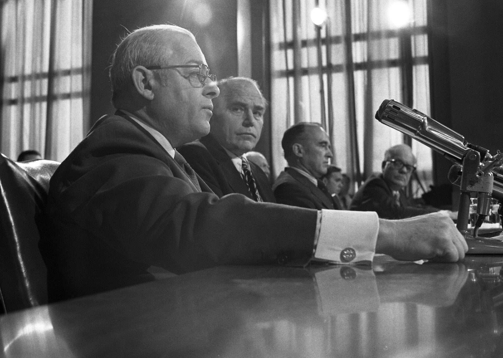 A black and white photos showing four men in suits sitting in front of a bench with a microphone