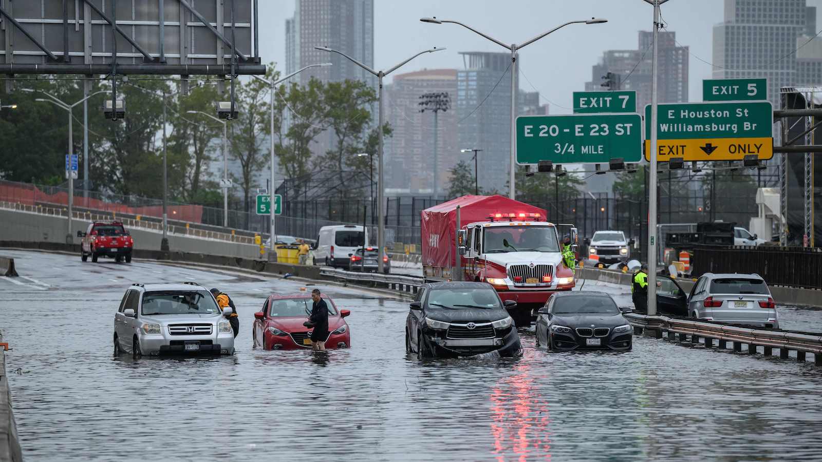 Four cars and a truck are half submerged in water on a highway with green exit signs in the background