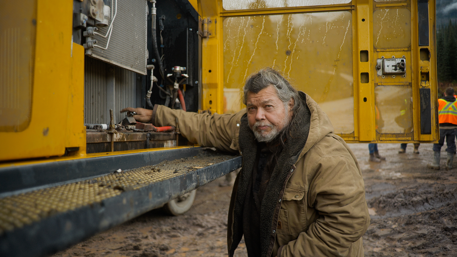 A man in a coat puts his arm on the back of a bulldozer