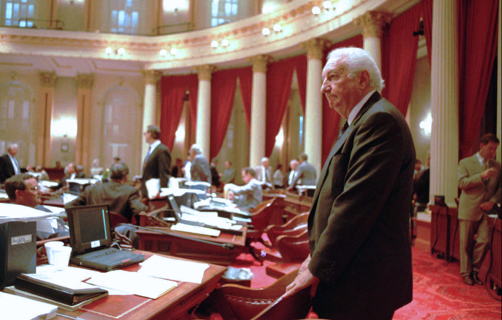 A man in a suit stands inside of a formal government chamber surrounded by desks with paper stacked on them