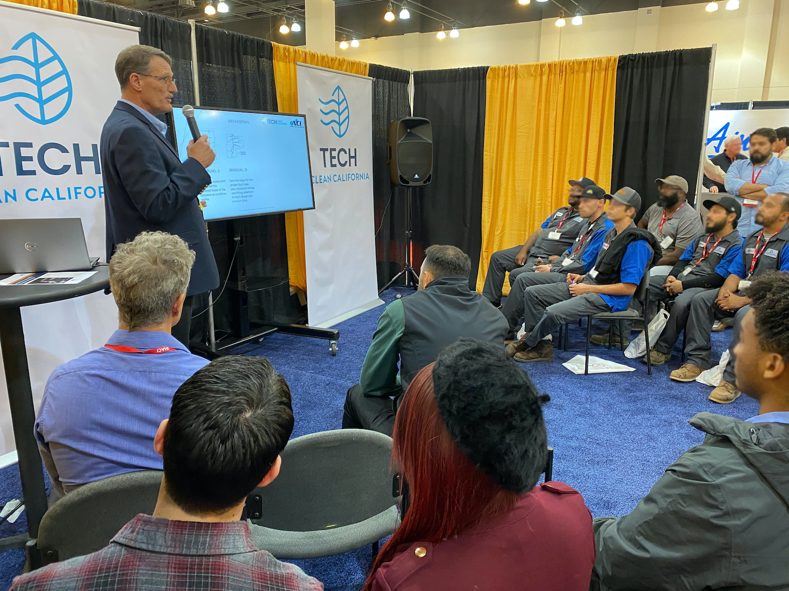 a group of people listens to a man in a suit talk on a microphone